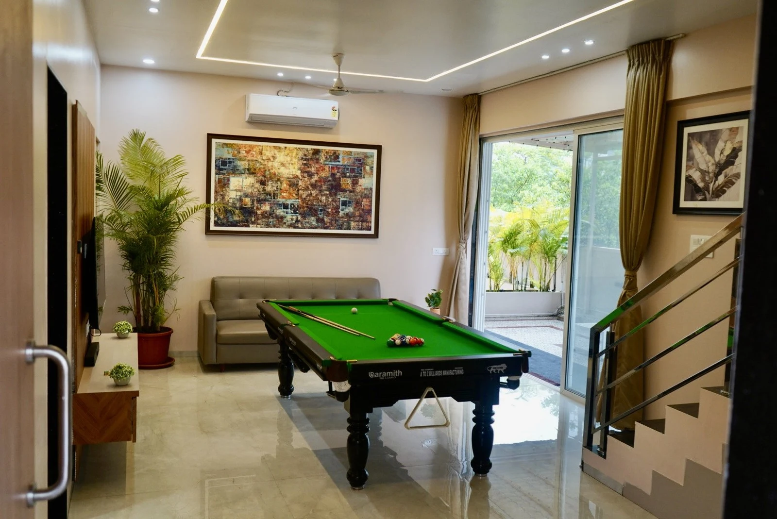 An uncovered green pool table in the living room, with a grey sofa, framed artwork and an open door to the pool deck.
