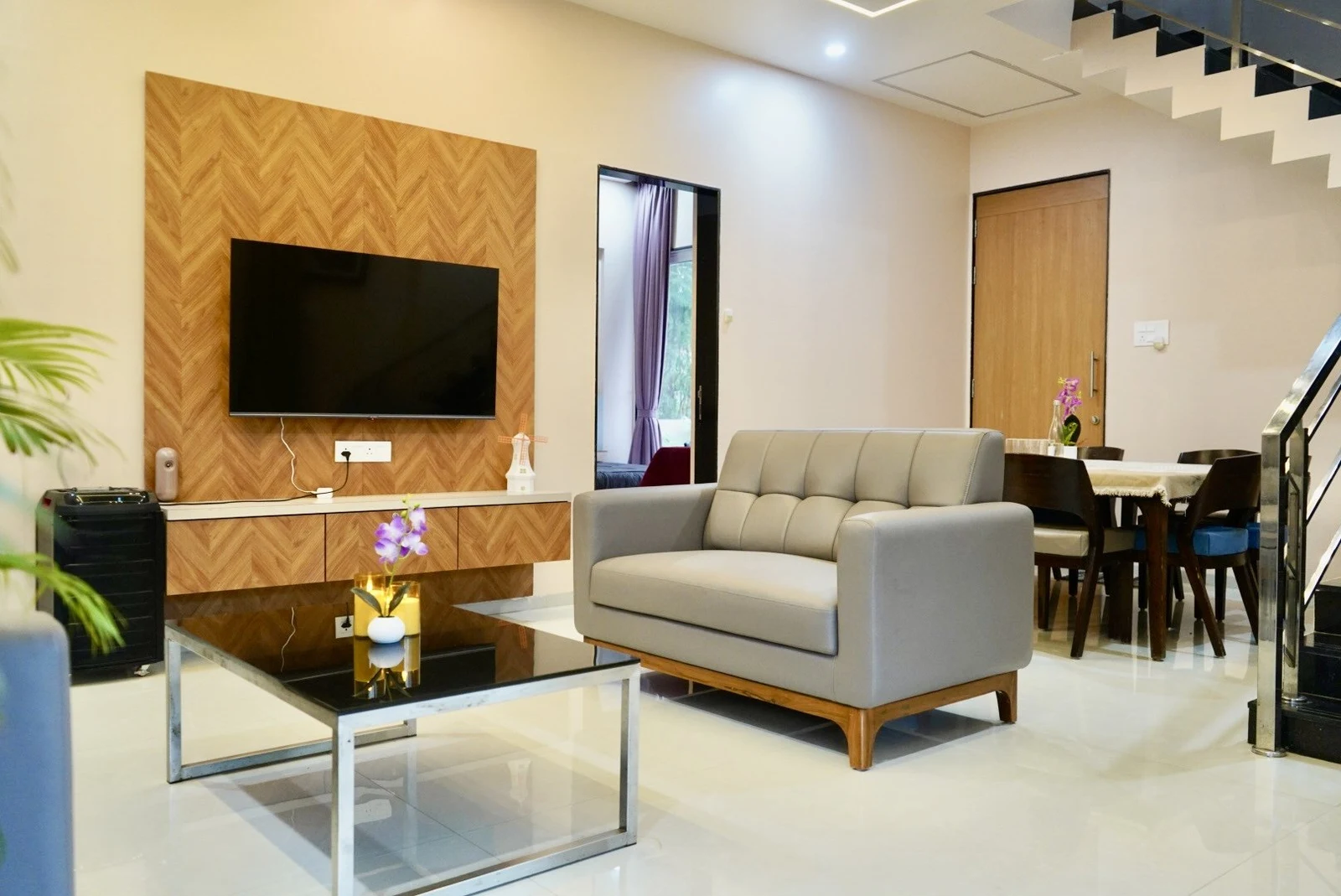 A bright lounge with grey seating, a glass coffee table, a wall-mounted television and the dining area behind it.